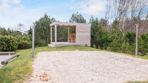 Une cabane en bois dotée d’un porche couvert, entourée d’une zone sablonneuse et d’une végétation luxuriante.