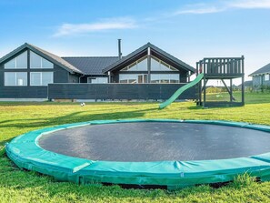 Ein Wohngebiet mit Trampolin im Vordergrund, einer Holzspielanlage und einem modernen Haus mit großen Fenstern und einem geneigten Dach.