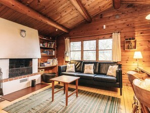 Interior acolhedor de uma cabana com lareira, um sofá, uma mesa e uma estante de livros.