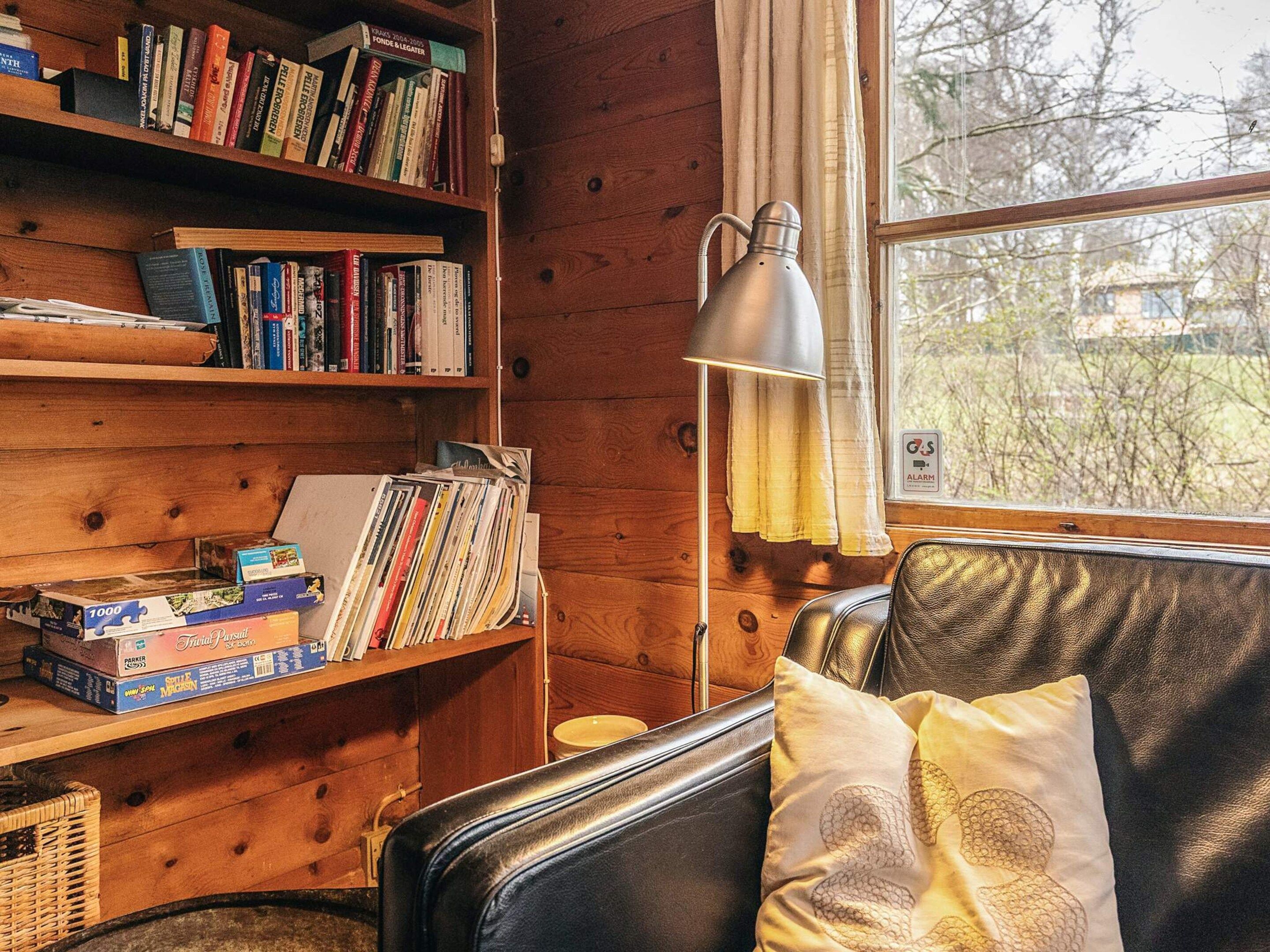 A cozy cabin interior with wooden walls, a bookshelf filled with books, a lamp, and a window with a view of trees.