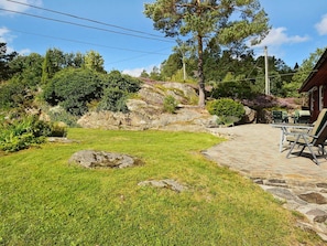 Um pátio de pedra com área de estar ao ar livre, rodeado por um gramado bem cuidado e diversos arbustos.