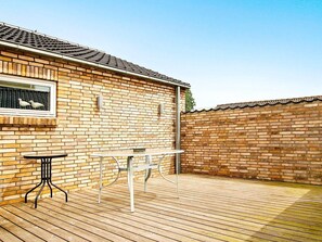 Une maison en briques avec un toit en tuiles, une terrasse en bois et deux tables en métal.