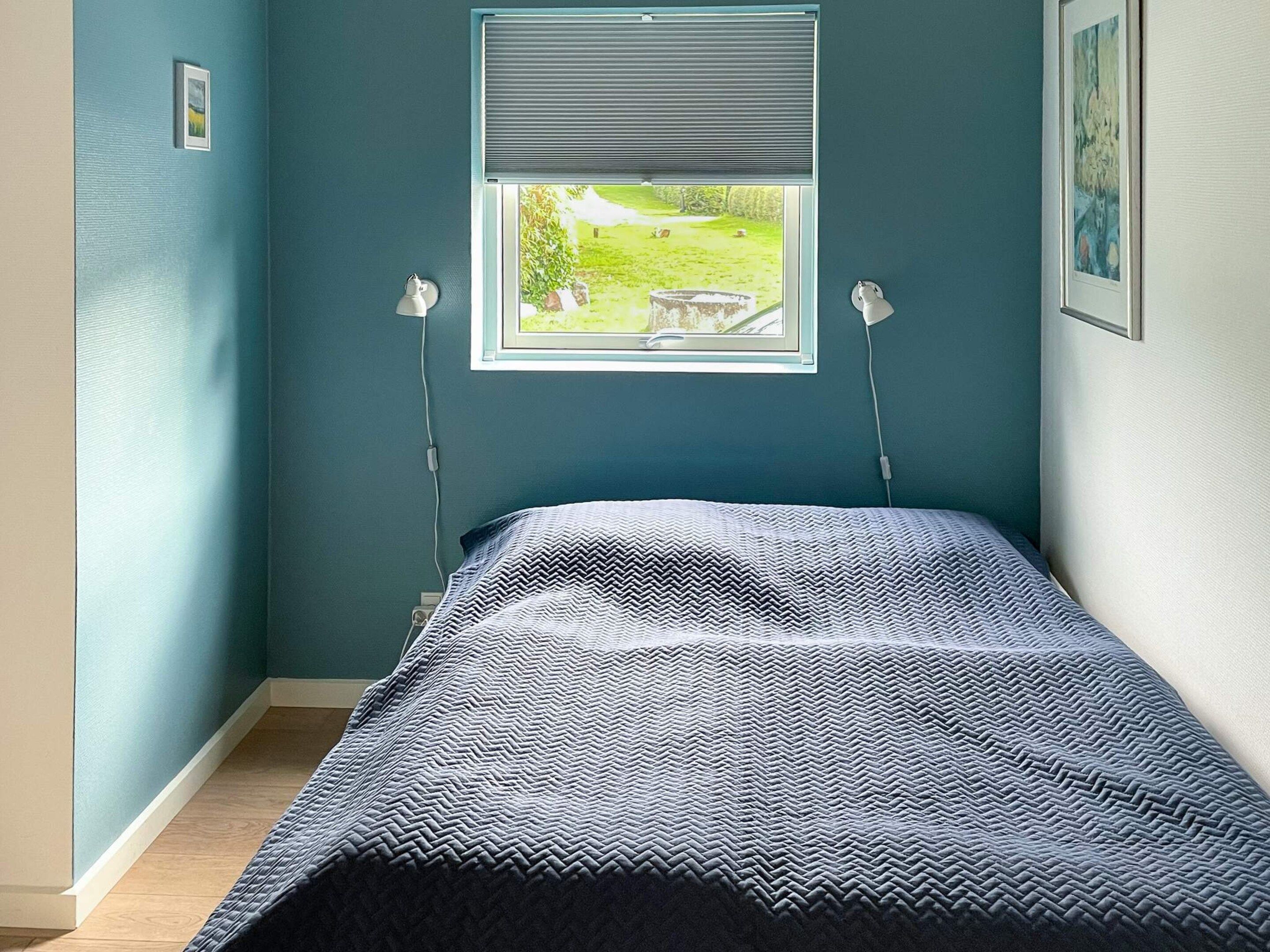 A bedroom with a bed, a window with a view of a garden, and two wall-mounted lamps.