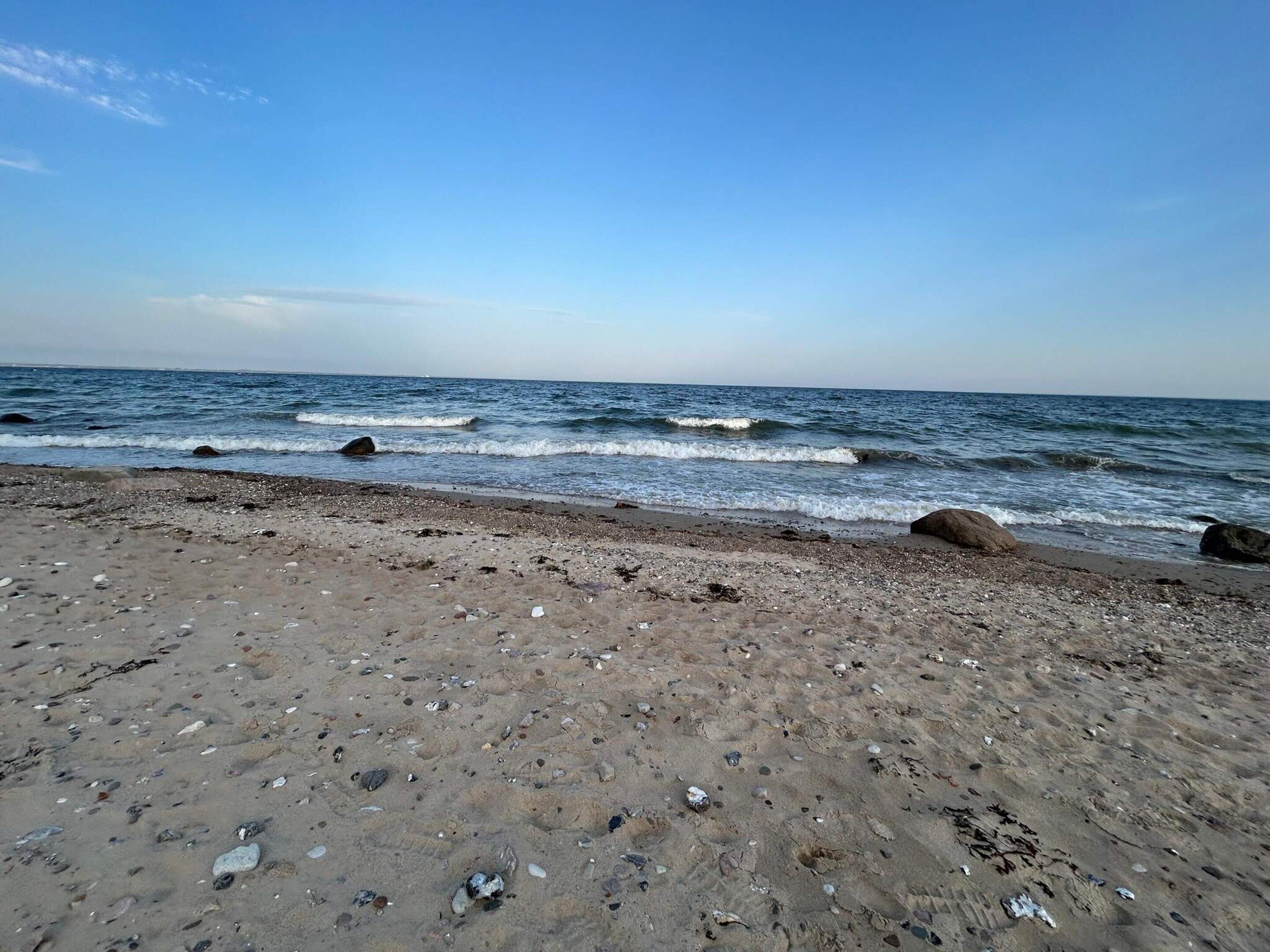 A beach with pebbles and rocks, a clear sky, and calm waves.
