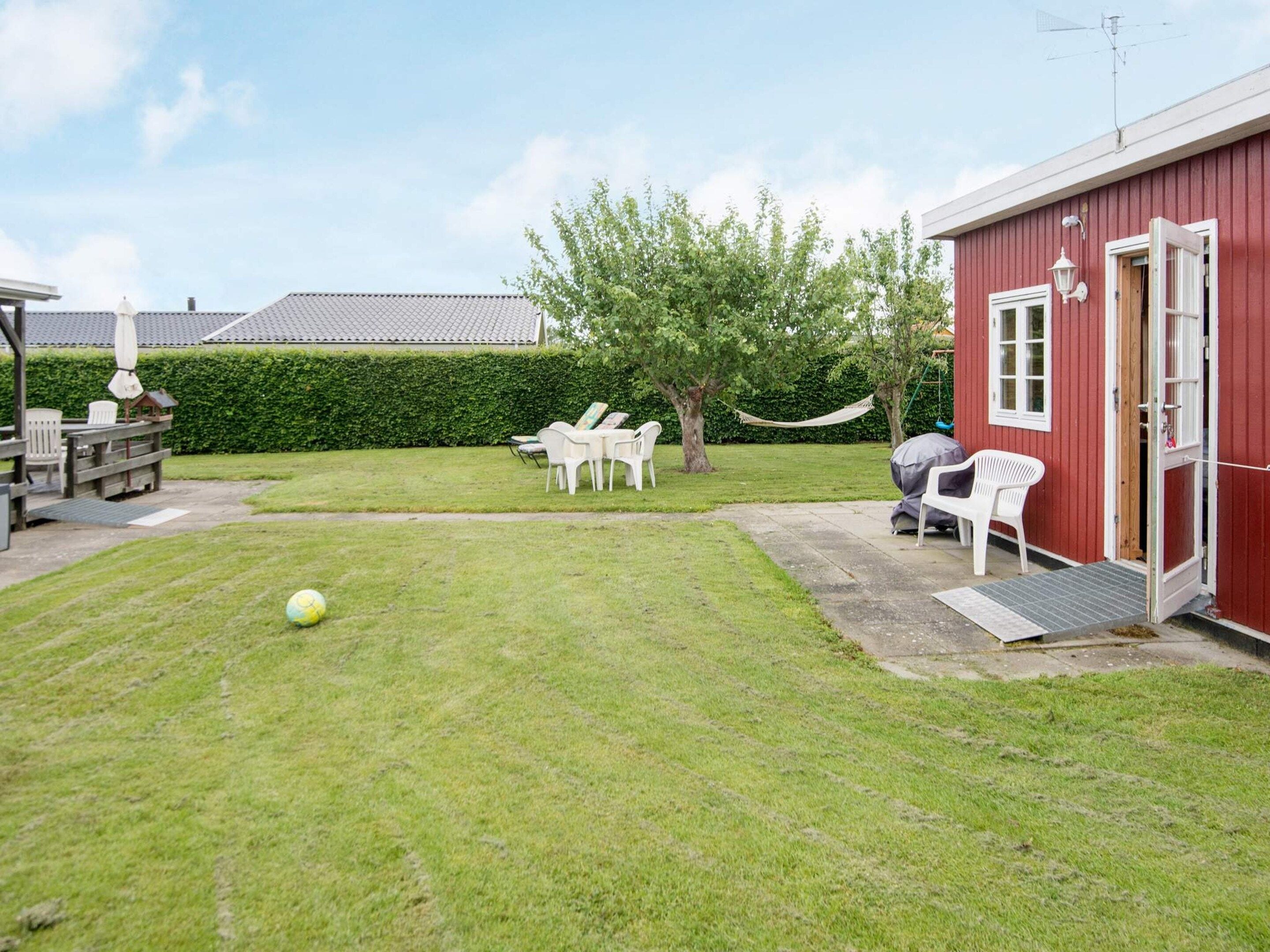 A red building with a door open, a grassy yard, white chairs, and a hammock.