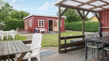 A covered outdoor seating area with a wooden deck, white plastic chairs, and a red building in the background.