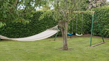 A hammock tied between two trees in a grassy area with a swing set and a hedge in the background.