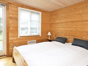 A wooden-paneled bedroom with a bed, a window with blinds, a bedside table with a lamp, and a vent.