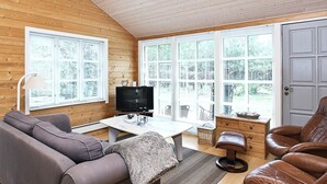 A cozy room with a wooden ceiling, a sofa, a leather chair, a TV, and large windows with blinds.