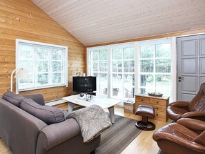A cozy room with a wooden ceiling, a sofa, a leather chair, a TV, and large windows with blinds.