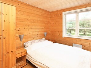 A wooden-paneled room with a bed, a nightstand, a lamp, and a window with a view of greenery.