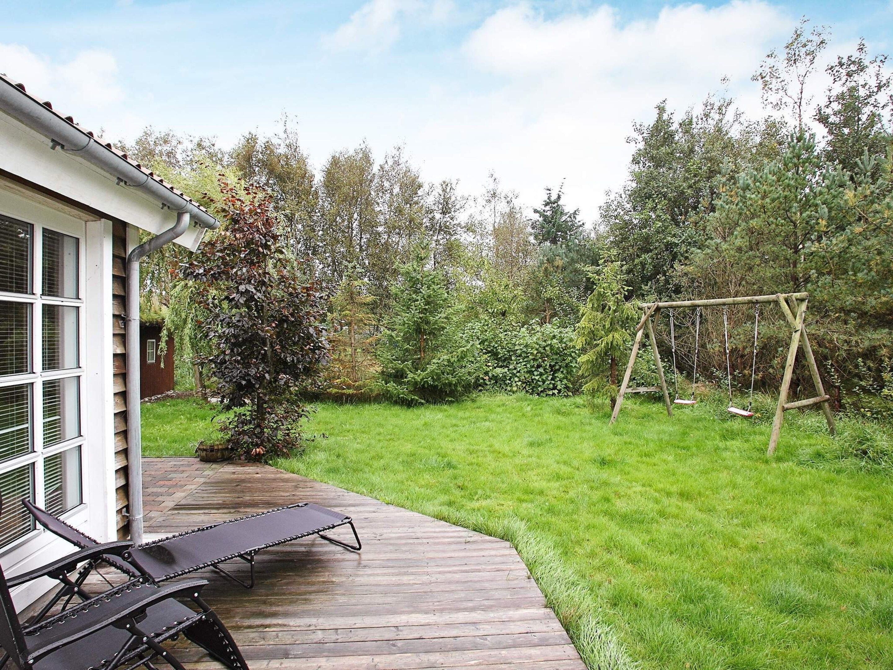 A wooden deck with a lounge chair, a swing set, and a garden with various trees and shrubs.