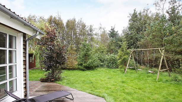 A wooden deck with a lounge chair, a swing set, and a garden with various trees and shrubs.