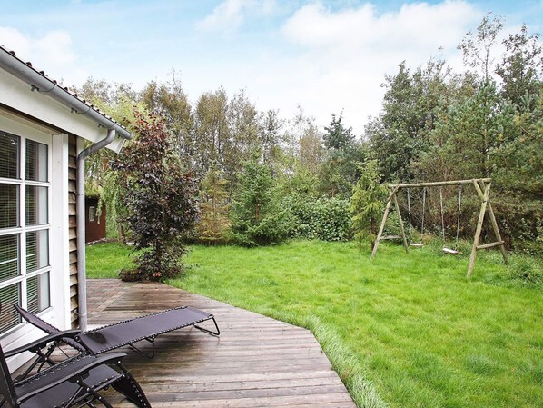 A wooden deck with a lounge chair, a swing set, and a garden with various trees and shrubs.