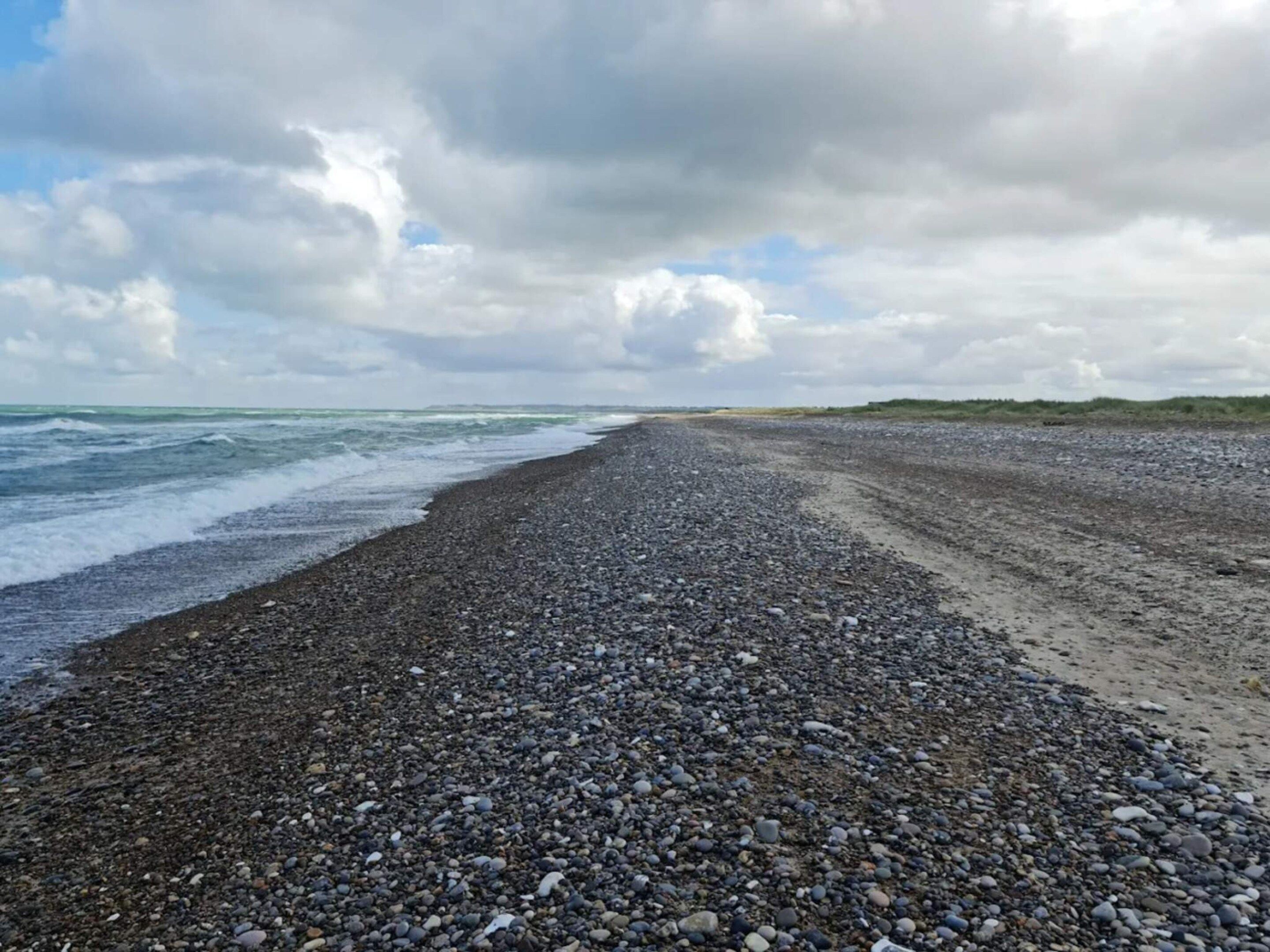 A pebbled beach with waves, a cloudy sky, and distant dunes.