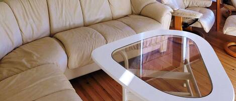 Un sofá de cuero color beige, una mesa de centro con cubierta de vidrio y una silla de madera en una habitación con piso de madera.