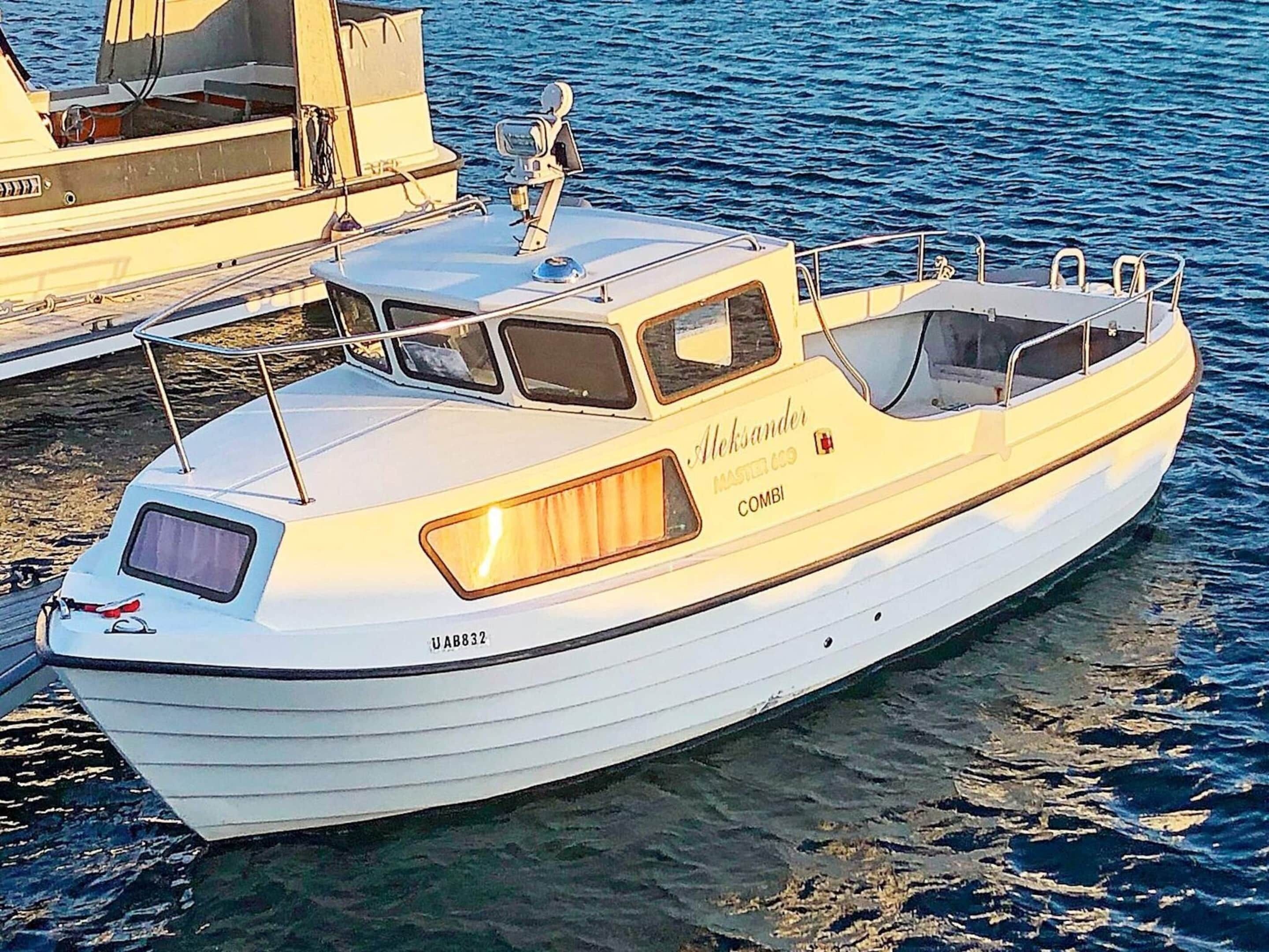 Um barco branco chamado 'Aksander' com cabine e janela, atracado em um porto.