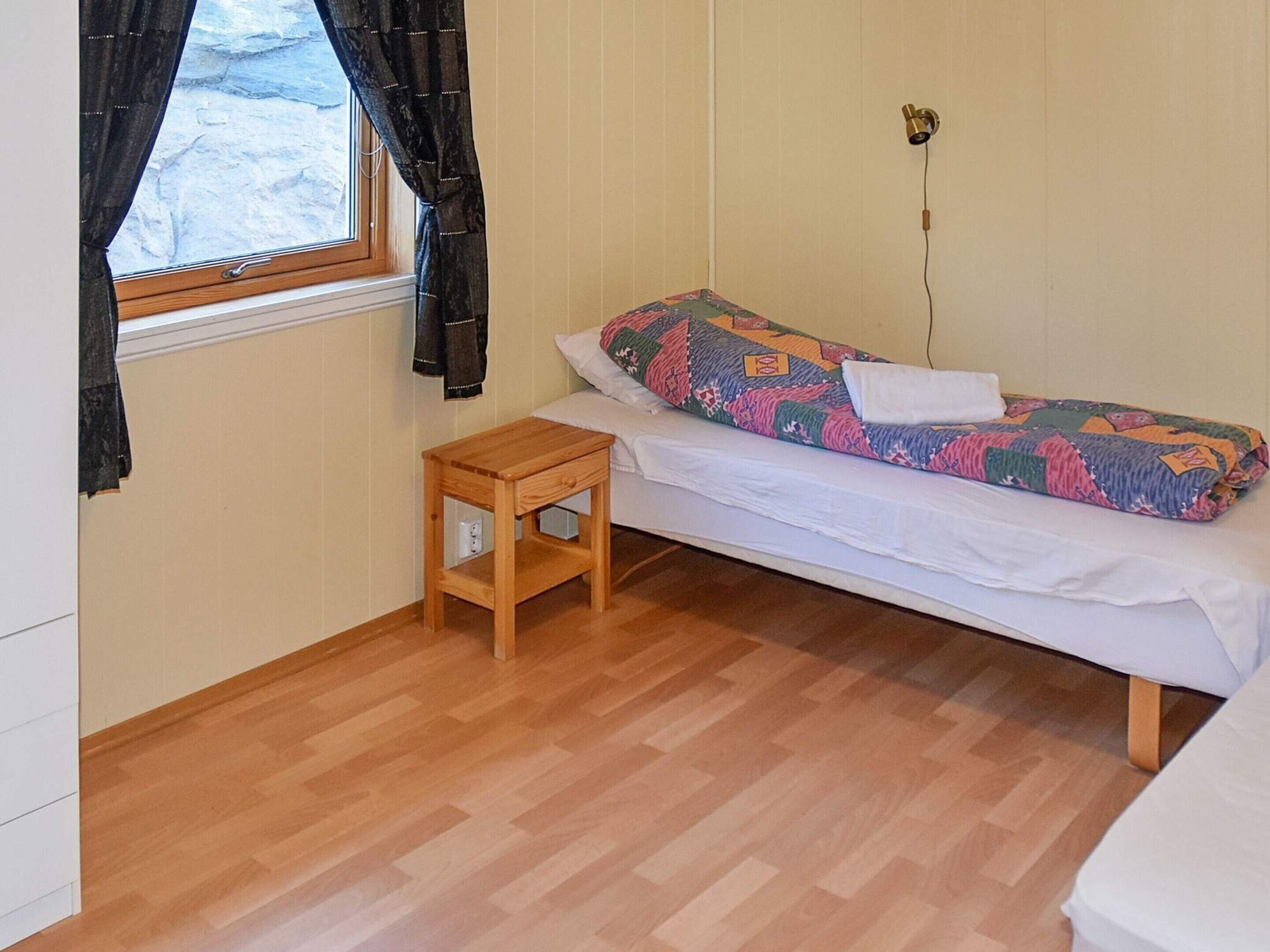 A small, single-bed room with a wooden floor, a small wooden nightstand, a window with curtains, and a wall-mounted lamp.