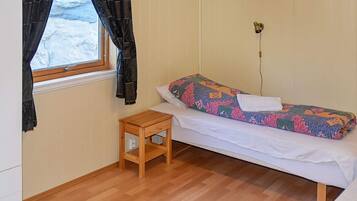 Un lit simple avec une couverture aux couleurs vives, une petite table de chevet en bois, une fenêtre avec des rideaux et une lampe fixée au mur.
