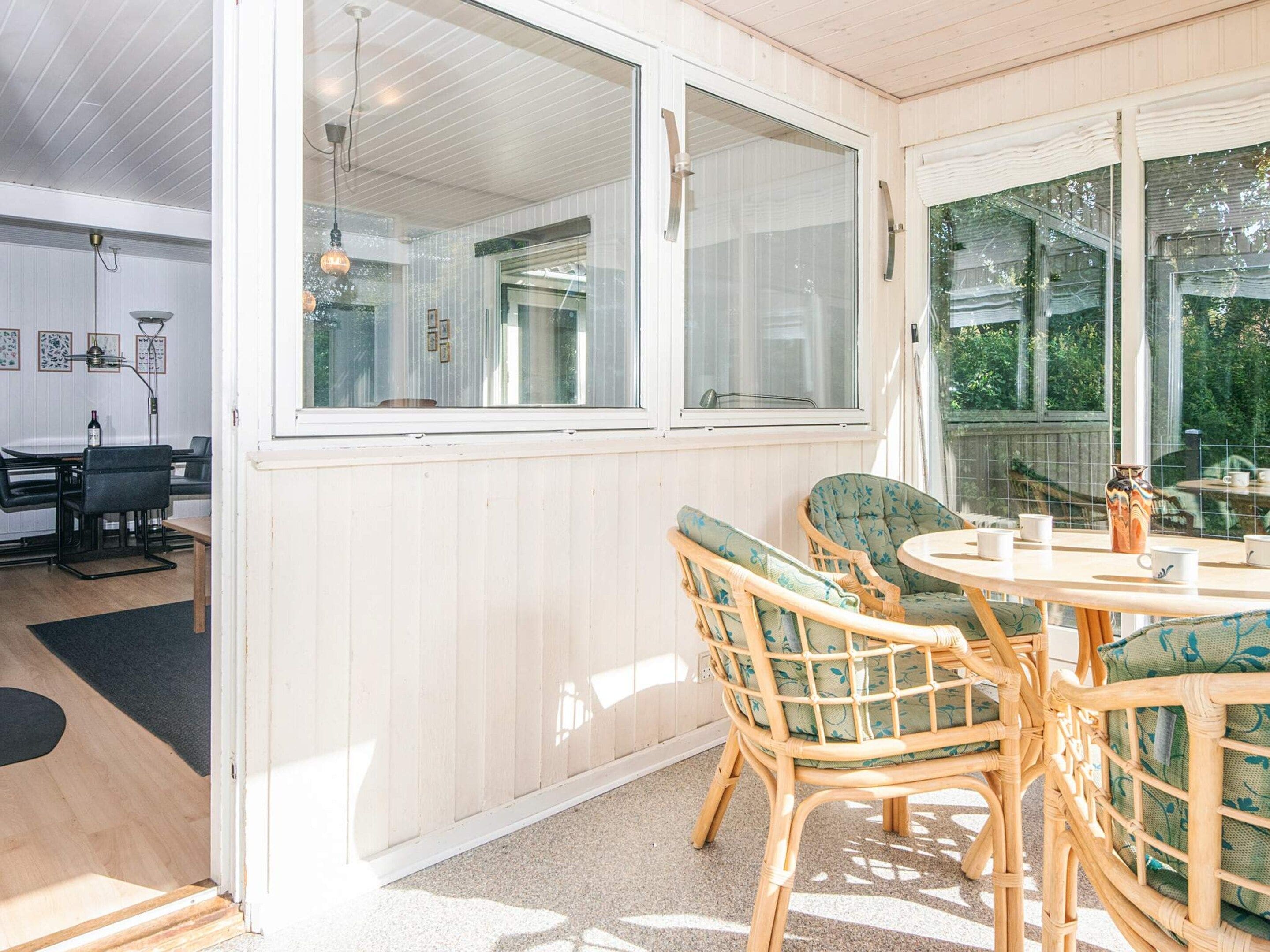A room with a dining area featuring wicker chairs and a round table, large windows with white frames, and a ceiling with wooden beams.
