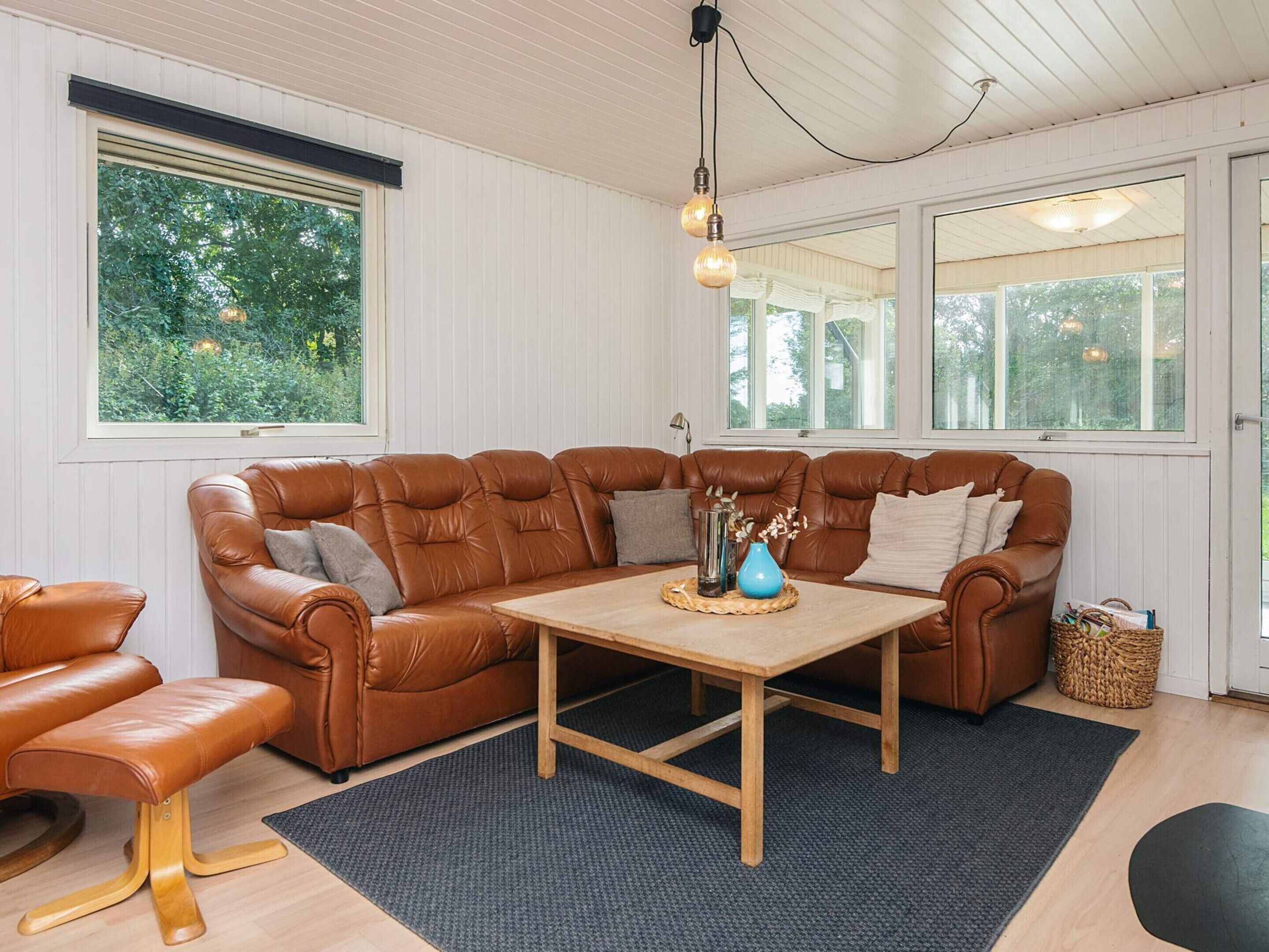 A cozy living room with a brown leather sofa, a wooden coffee table, and a large window with a view of greenery.