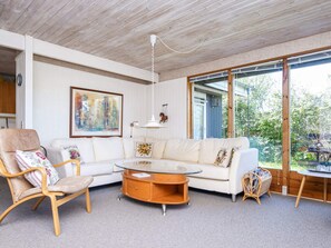 A modern living room with a white sofa, a wooden chair, a glass coffee table, and a large window offering a view of greenery. - 6 Person Holiday Home in Glesborg-by Traum (Glesborg)