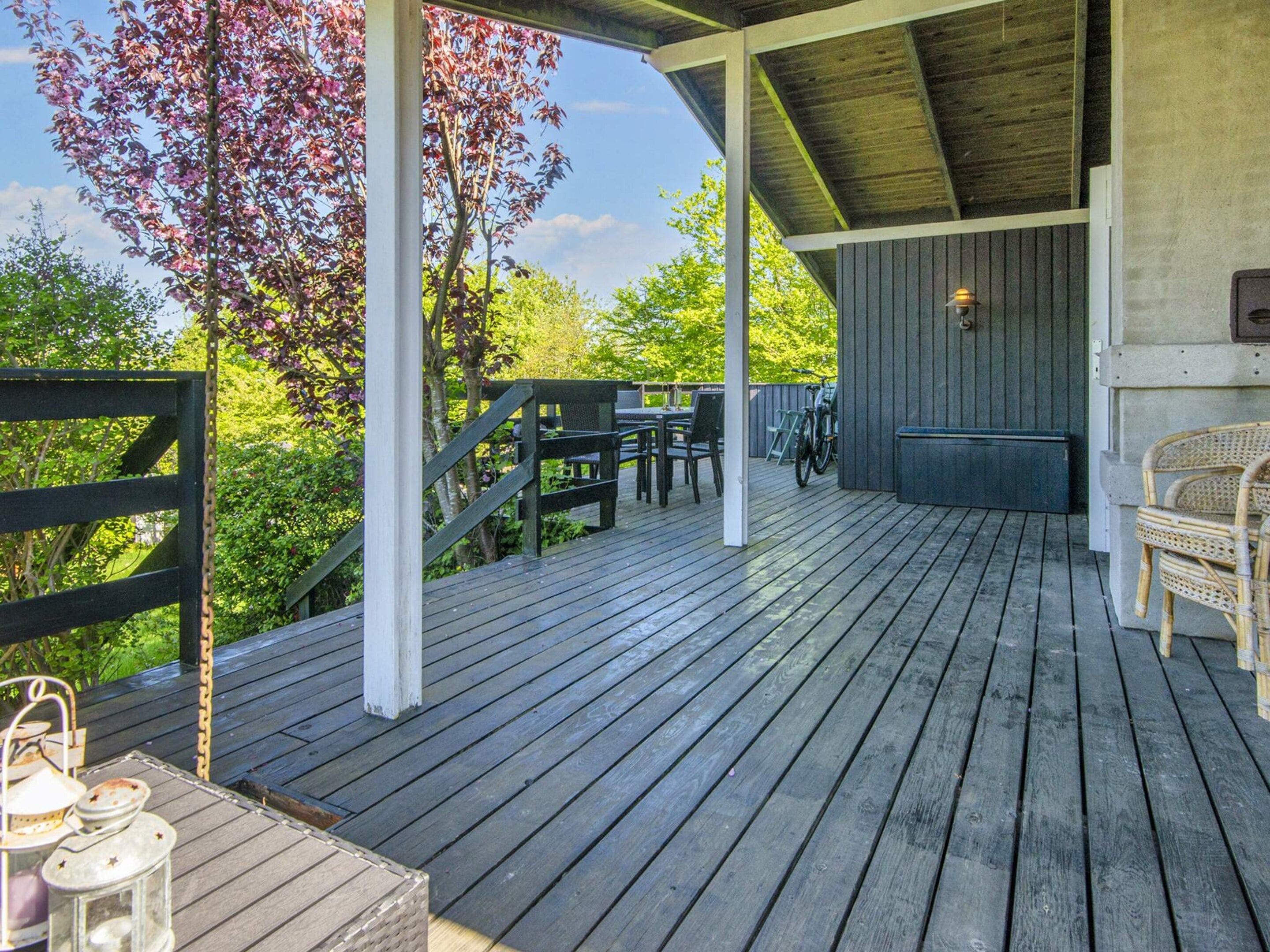 Une terrasse extérieure couverte avec un plancher en bois, un espace salon avec des chaises et des tables, et un vélo garé au fond.