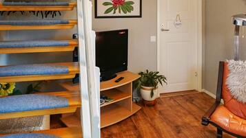 A staircase with blue carpeted steps leading to a landing, a television on a wooden stand, a potted plant, and a chair with a fluffy pillow.