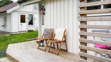 A wooden deck with two chairs and a small table, a white building with a door and windows, and a wooden fence.