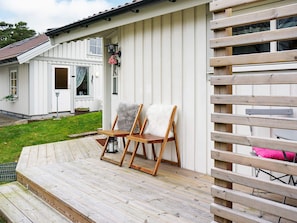 A wooden deck with two chairs and a small table, a white building with a door and windows, and a wooden fence.