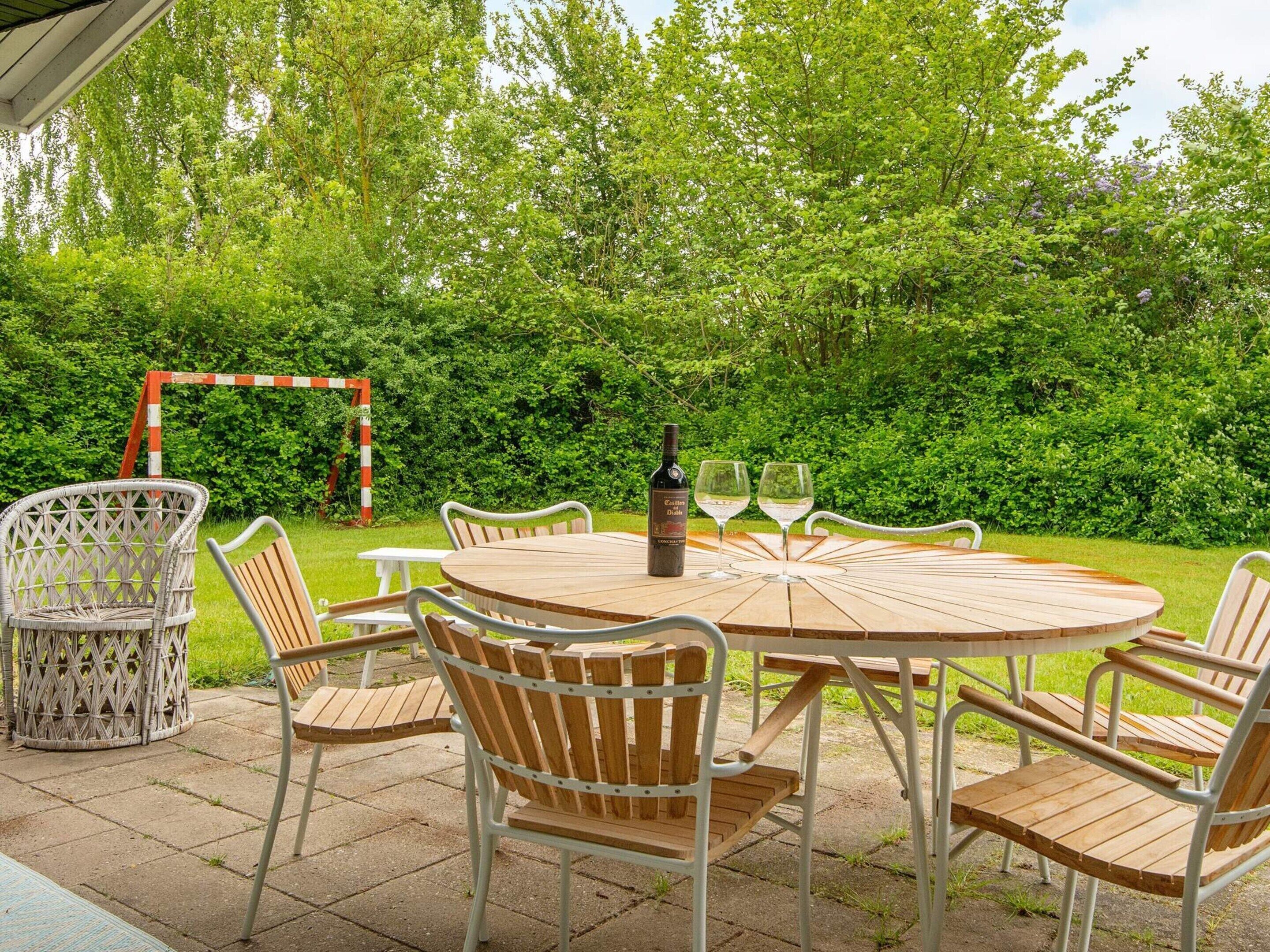 An outdoor patio with a round wooden table and six chairs, surrounded by lush greenery.