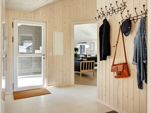 Un pasillo con una puerta de madera, un perchero con sombreros y bolsos, y una vista de una sala de estar con sofá y sillones.