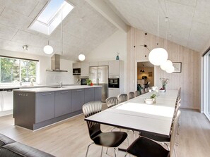 A modern kitchen with a central island, a dining area with a long table and chairs, and a view of the outdoors through large windows. - 5 Star Holiday Home in Frederiksvaerk (Frederiksvaerk)