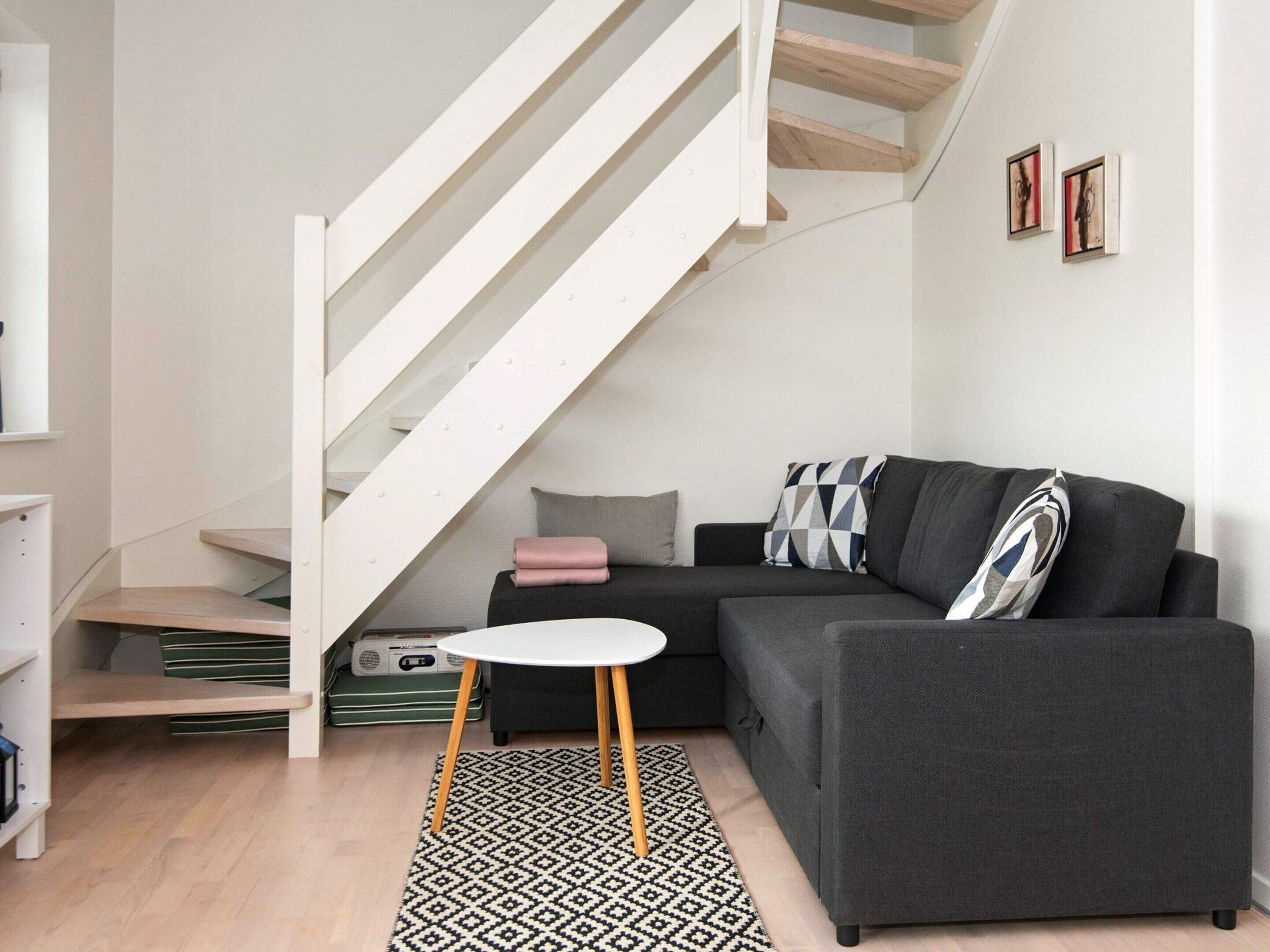 Un salon moderne avec un escalier, un canapé gris, une petite table ronde et un tapis à motifs.