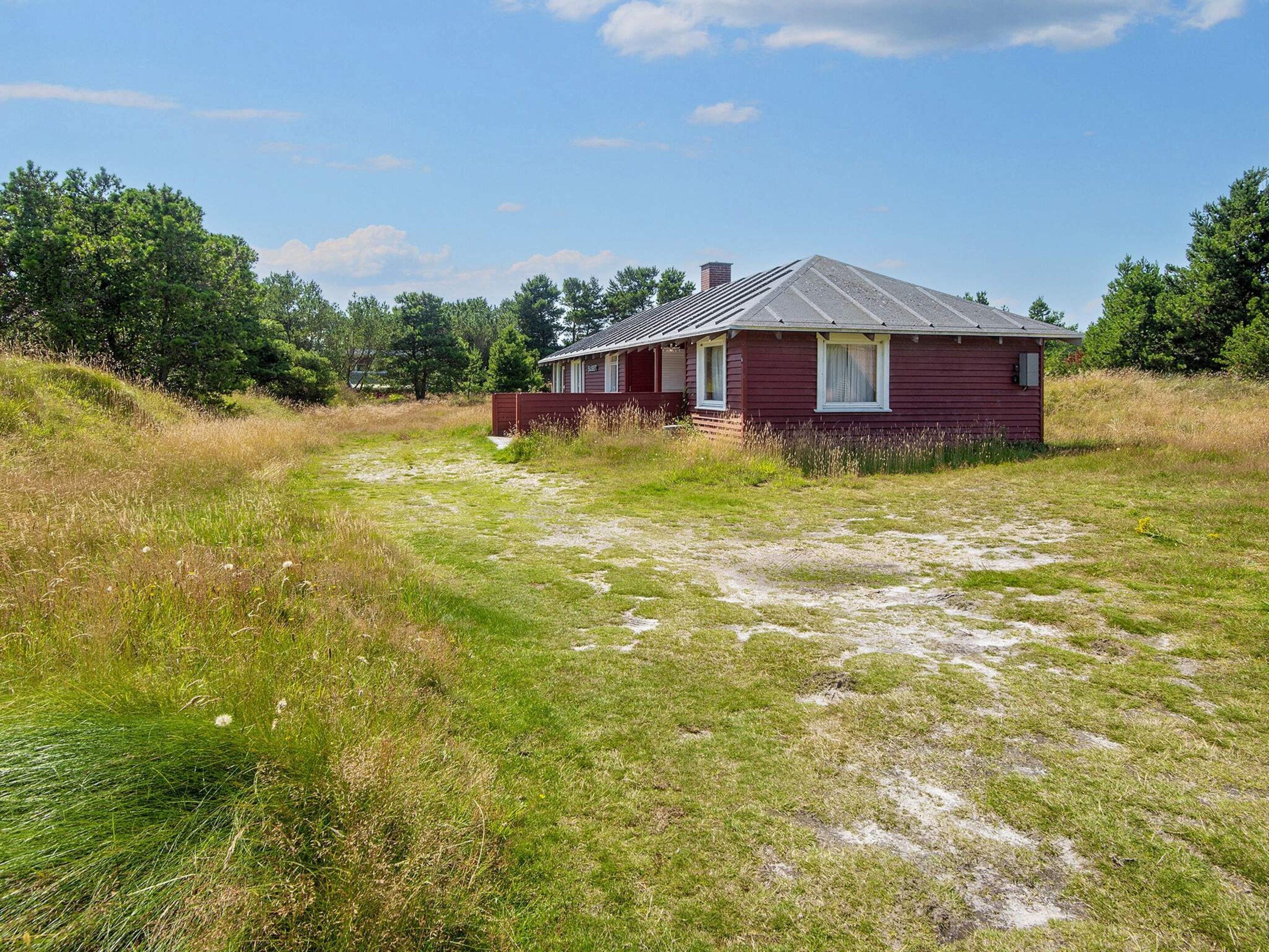 A single-story house with a sloped roof, surrounded by a grassy field with trees in the background.