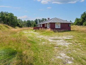 A single-story house with a sloped roof, surrounded by a grassy field with trees in the background.