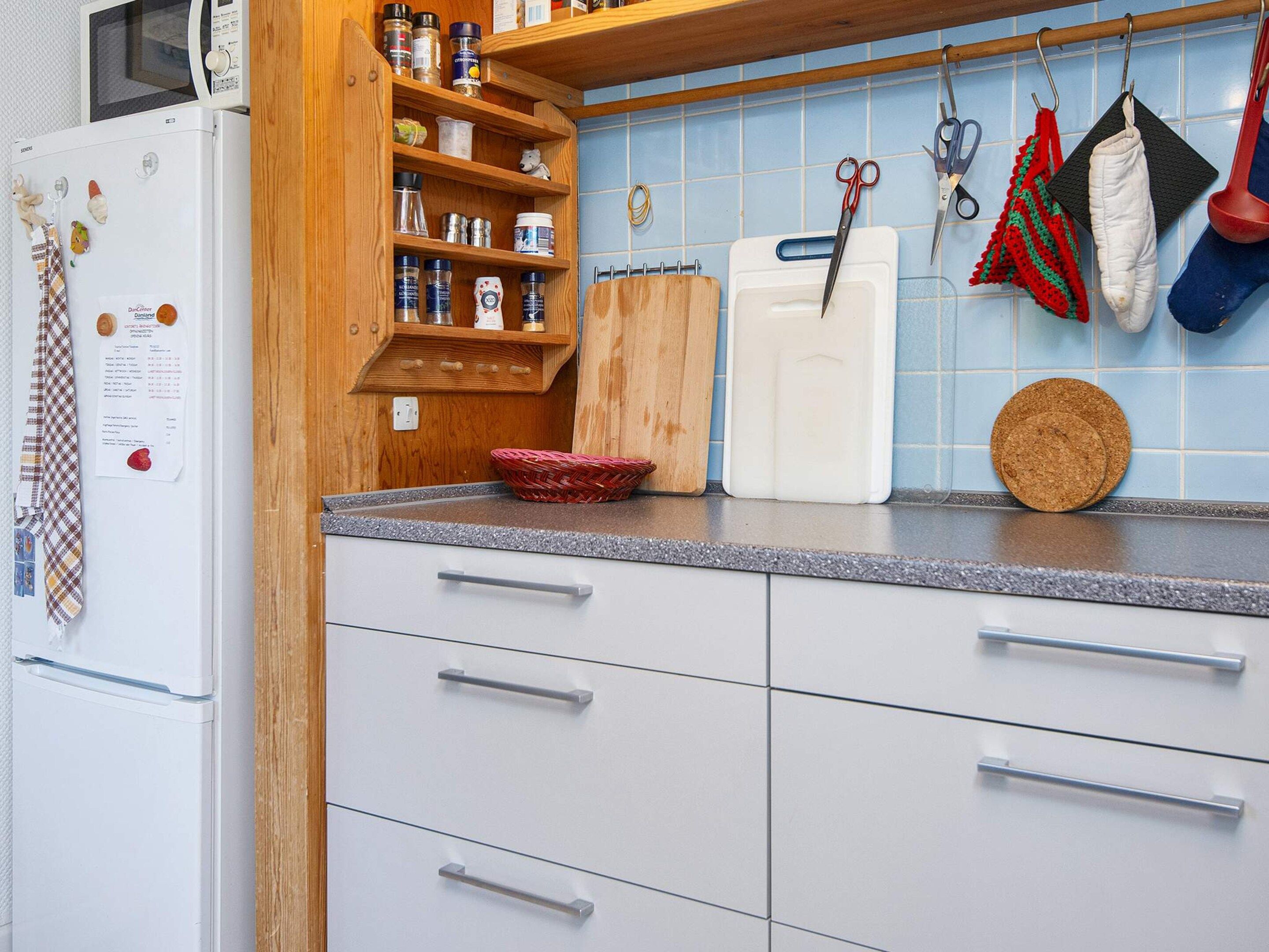 A kitchen with a refrigerator, wooden shelves, a cutting board, a large white container, and kitchen utensils.