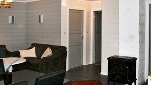 Ein Wohnzimmer mit einem dunkelgrünen Sofa, einem kleinen Tisch und einem roten Teppich. Auf dem Boden steht ein schwarzes Radio, und im Hintergrund ist eine weiße Tür zu sehen.
