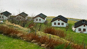 Eine Reihung weißer Häuser mit rotem Zierstreifen und dunklen Dächern, auf einem grasbewachsen Hügel mit Blick auf ein Gewässer.