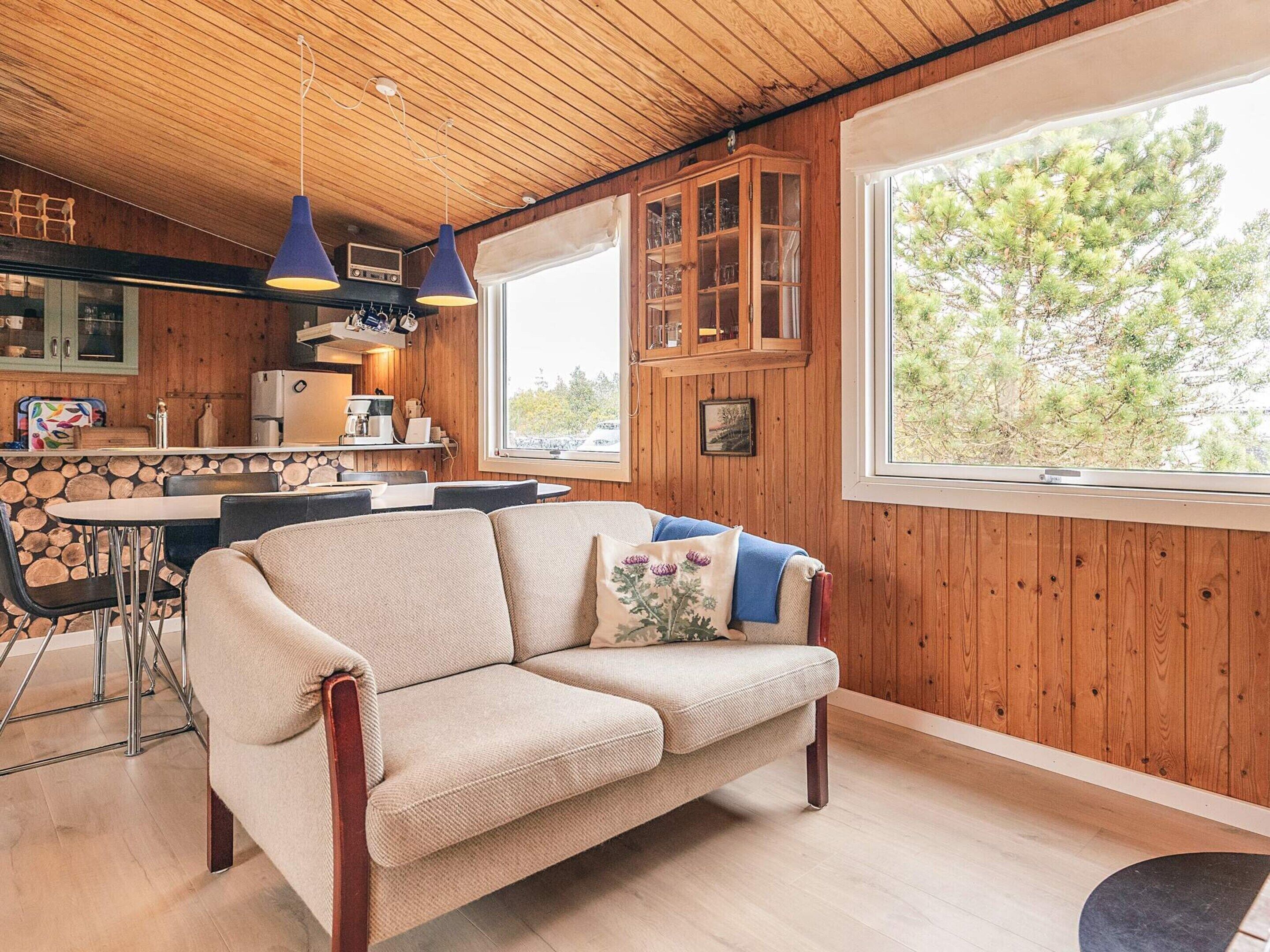 A cozy cabin interior with a wooden ceiling, a sofa with cushions, a dining table with chairs, and a large window with a view of trees.