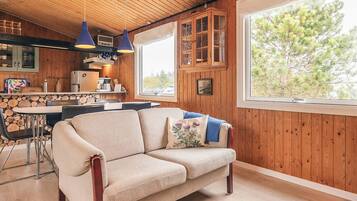 A cozy cabin interior with a wooden ceiling, a sofa with cushions, a dining table with chairs, and a large window with a view of trees.