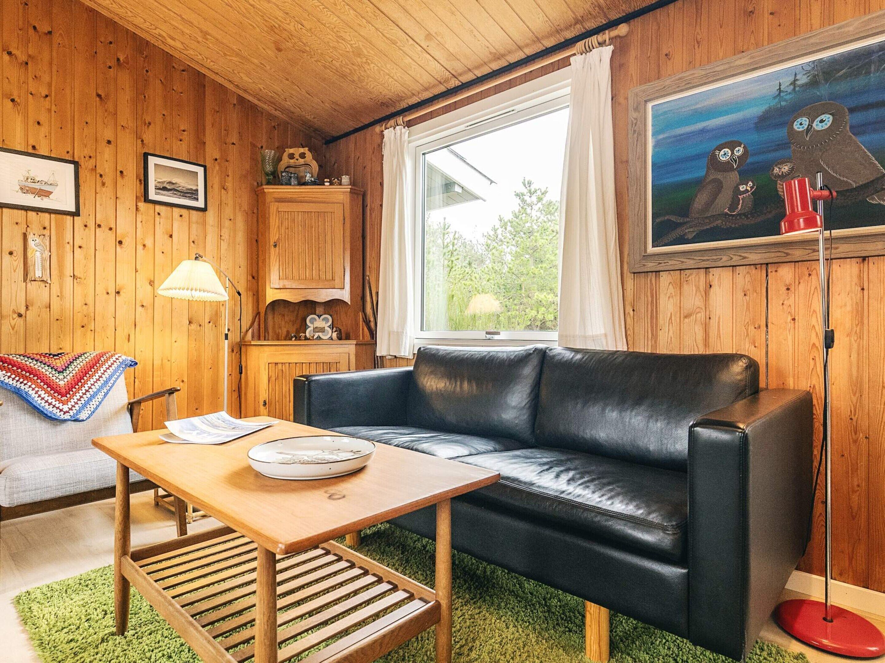 Ein gemütliches Zimmer mit einem schwarzen Ledersofa, einem Holztisch und einem grünen Teppich. Ein Fenster mit weißen Vorhängen und ein Gemälde von Eulen an der Wand.