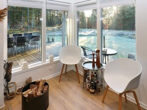 Une pièce chaleureuse avec une grande fenêtre donnant sur une terrasse et une forêt. On y trouve deux chaises blanches, une petite table ronde avec des bouteilles, et une cheminée avec des bûches.