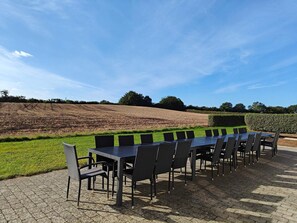 Een buiten eetgedeelte met een lange tafel en stoelen, gelegen op een verhard oppervlak met uitzicht op een bewerkt veld en een haag op de achtergrond.