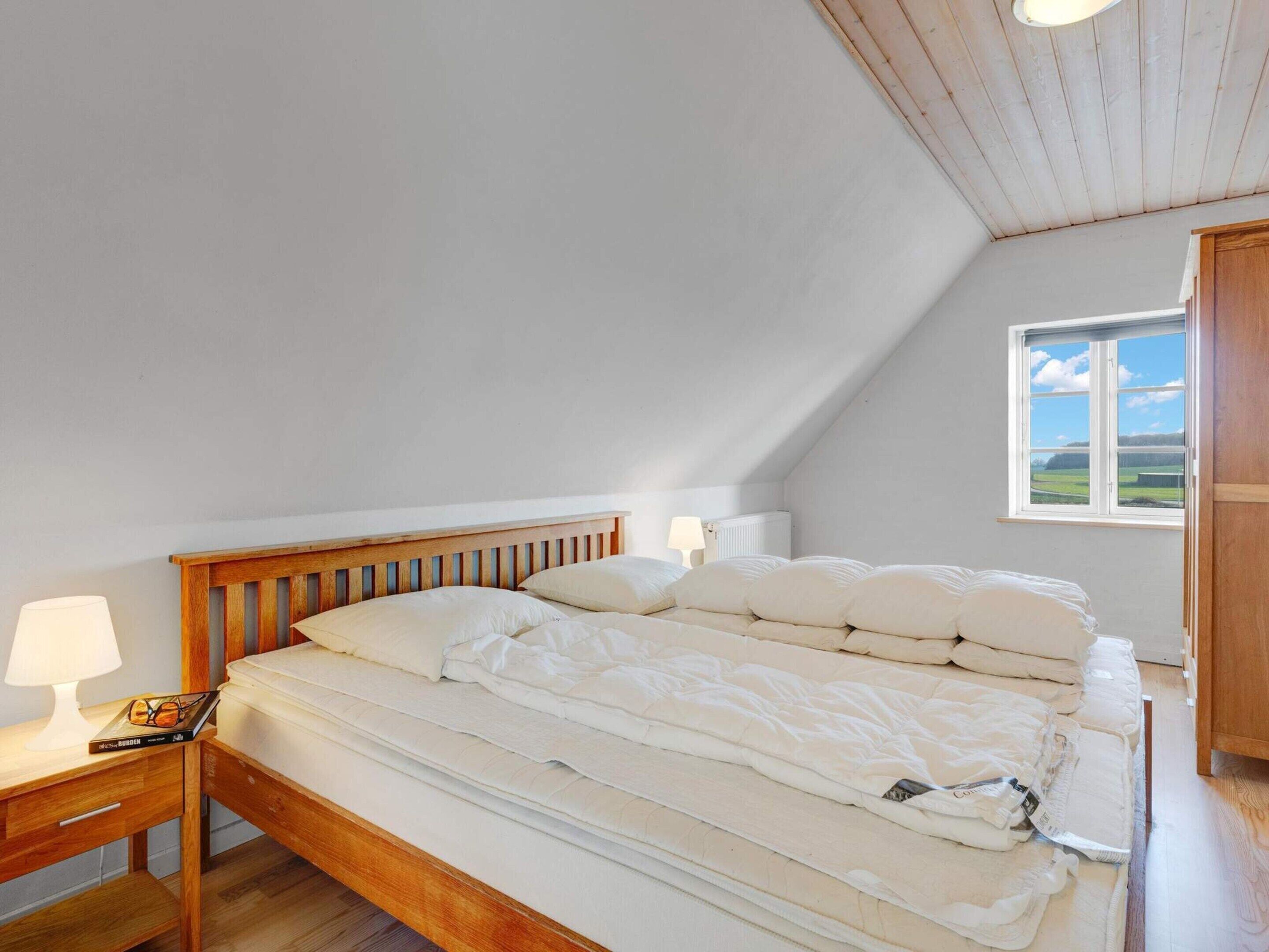 Een slaapkamer met een houten bed, wit beddengoed, een nachtkastje met lamp en een raam met uitzicht op het landschap.