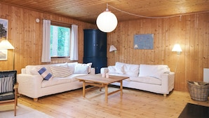 En stuga med träpanel på insidan, två vita soffor, ett träbord, ett stort fönster med gardiner och en vägglampa.