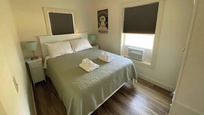 A bedroom with a bed, two bedside tables, a nightstand, a window with blinds, and a poster on the wall.