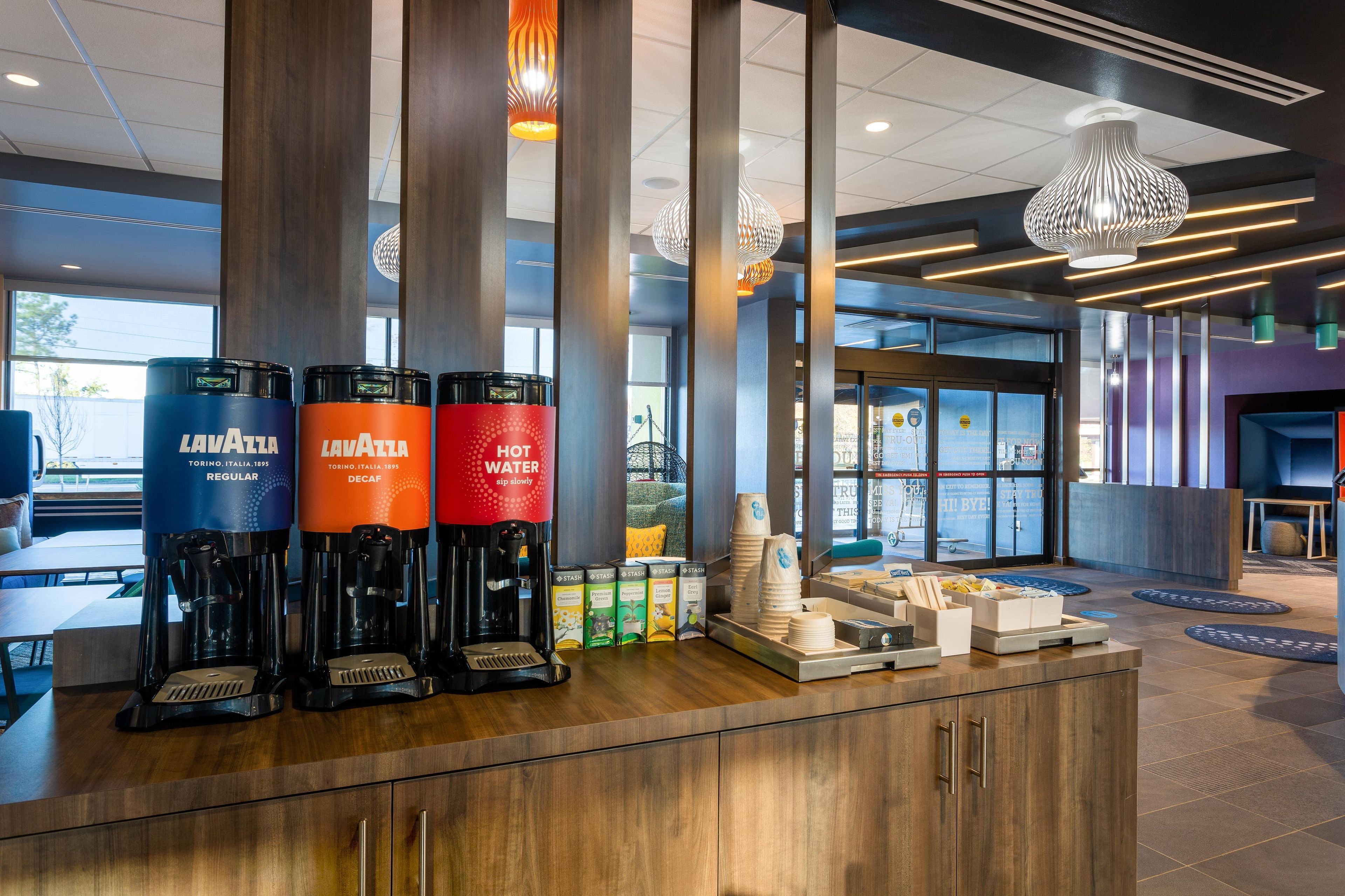 Zona de recepción de hotel con una estación de café que incluye máquinas de espresso Lavazza, un área de descanso con mesas y vistas al exterior a través de amplios ventanales.