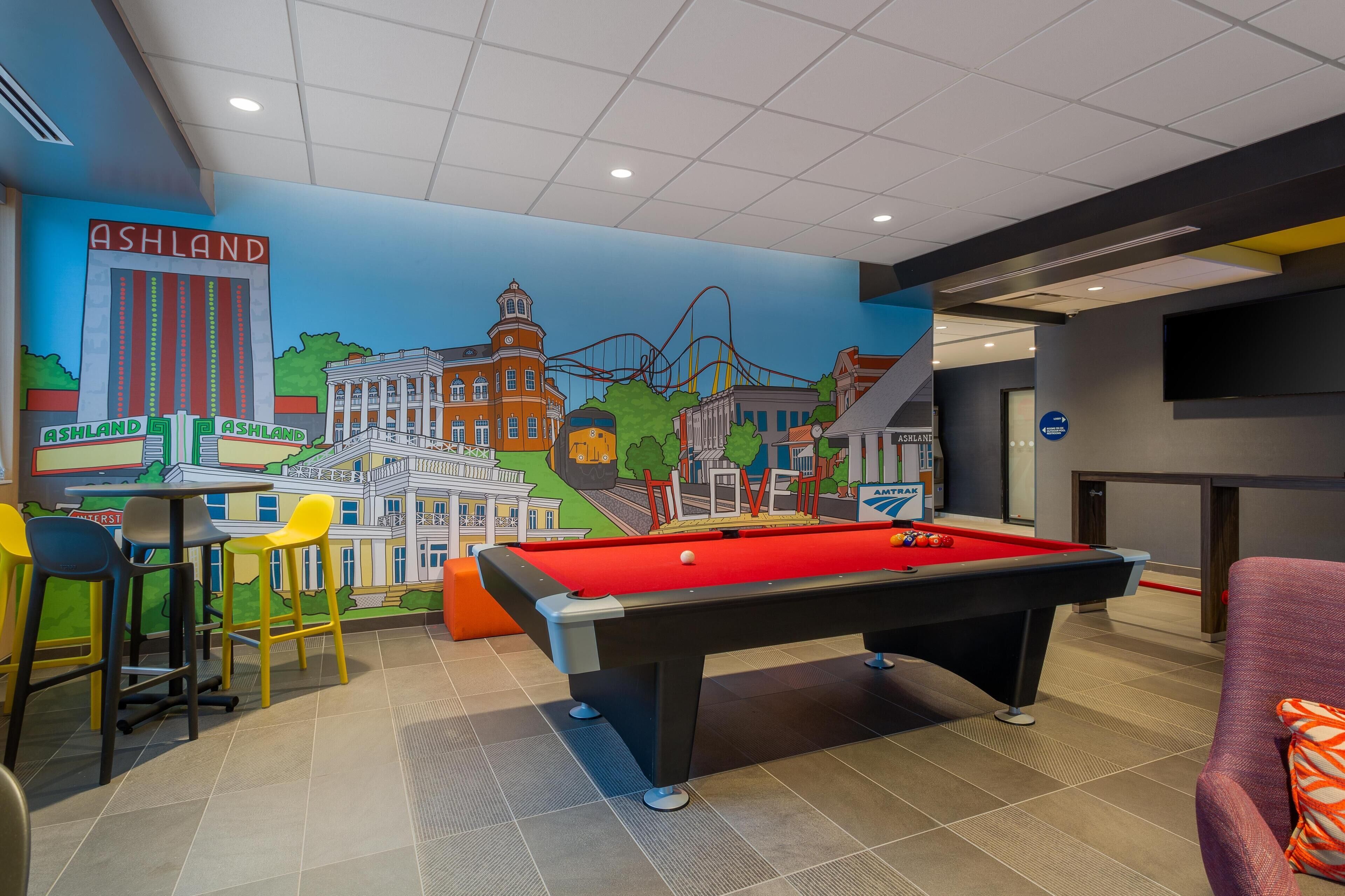 Een pooltafel met een rode viltbekleding, een bar met krukjes, en een muurschildering waarop een stadsgezicht te zien is, met de tekst 'ASHLAND' op de muur.