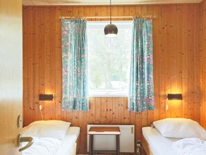 A room with two beds, wooden walls, a window with curtains, and a table.